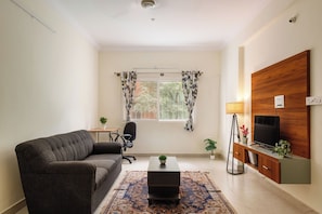 Un salón con un sofá, una mesa de centro, un televisor sobre un mueble de madera, una mesita con una lámpara y una ventana con cortinas.