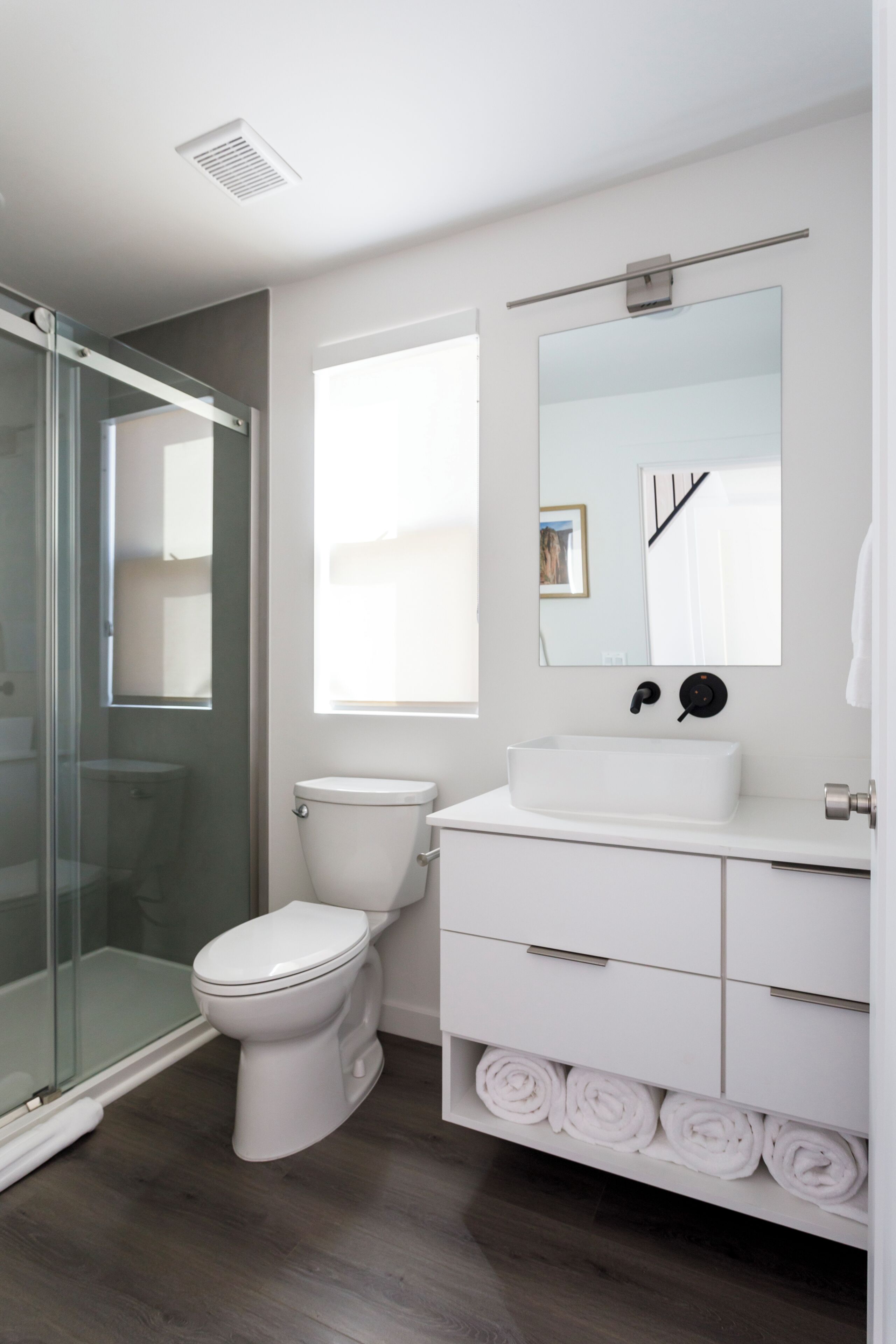 A modern bathroom with a glass shower enclosure, a white toilet, a sink with a mirror above, and neatly rolled towels on a shelf.