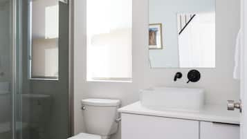 A modern bathroom with a glass shower enclosure, a white toilet, a sink with a mirror above, and neatly rolled towels on a shelf.