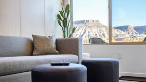 Ein modernes Wohnzimmer mit einer Couch, Poufs, einer Fernbedienung und einem großen Fenster mit Blick auf eine Berglandschaft.