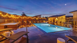Une piscine dotée de chaises longues et d’un jacuzzi, le tout entouré de bâtiments à l’éclairage chaleureux.