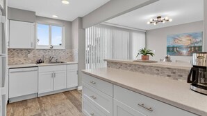A modern kitchen with white cabinets, a tiled backsplash, and a large window. - Ocean Breeze Condo | Ocean Views | Steps To Beach | Pool | Flagler/Ormond Beach (Ormond Beach)
