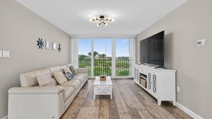 A living room with a beige sofa, a white coffee table, and a TV on a white cabinet. - Ocean Breeze Condo | Ocean Views | Steps To Beach | Pool | Flagler/Ormond Beach (Ormond Beach)