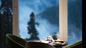 A dining area with a table set for two, a view of a snowy landscape through large windows, and a green chair. - EAFR Hotel (Nathia Gali)
