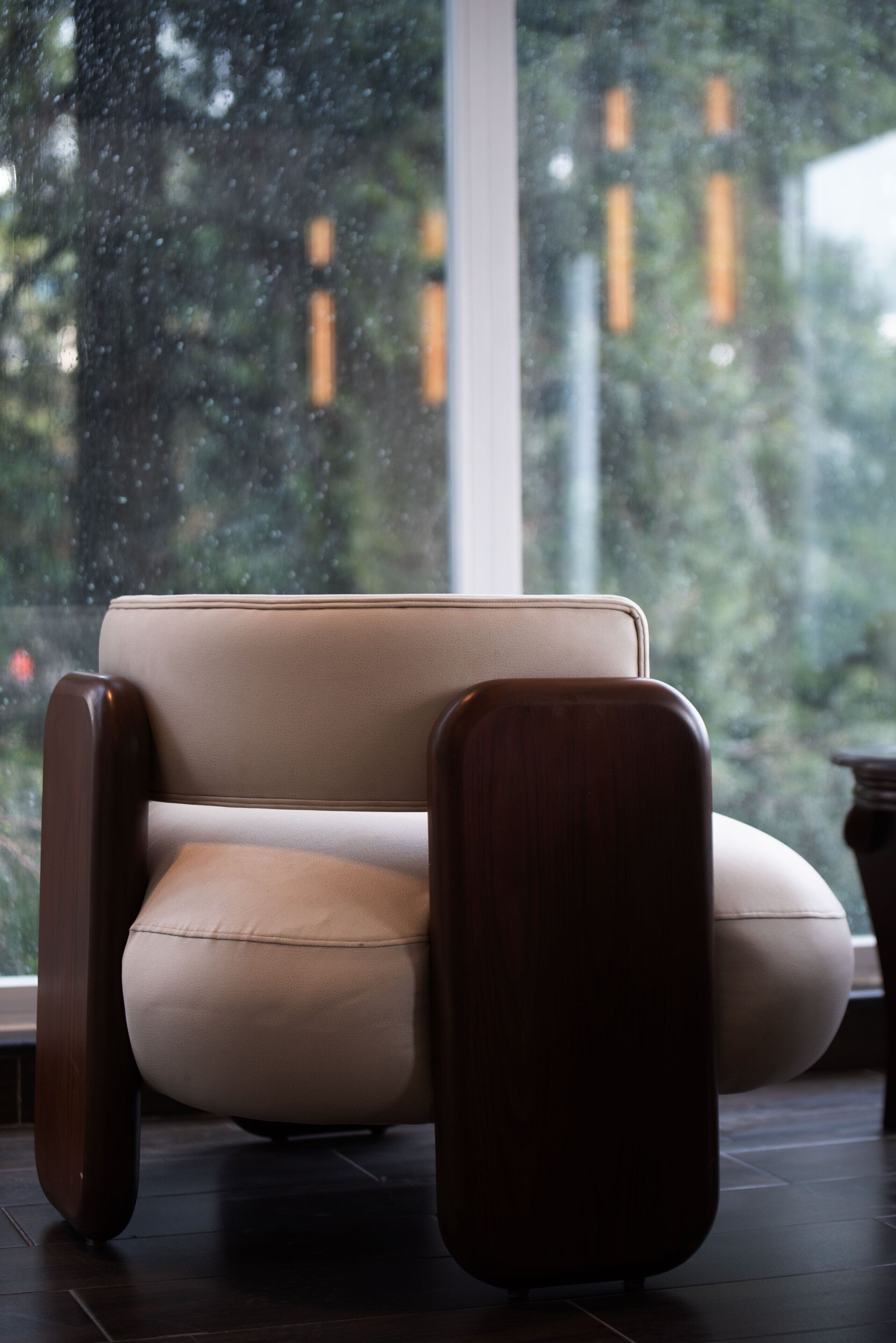 Una silla moderna con estructura de madera y asiento acolchado, ubicada frente a una ventana con vistas a árboles y edificios.