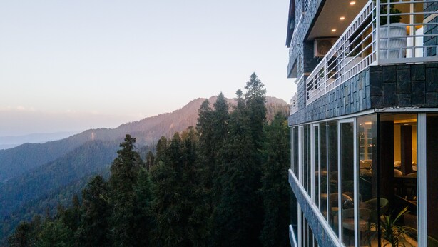 A modern building with a glass facade overlooking a forested mountain range. - EAFR Hotel (Nathia Gali)