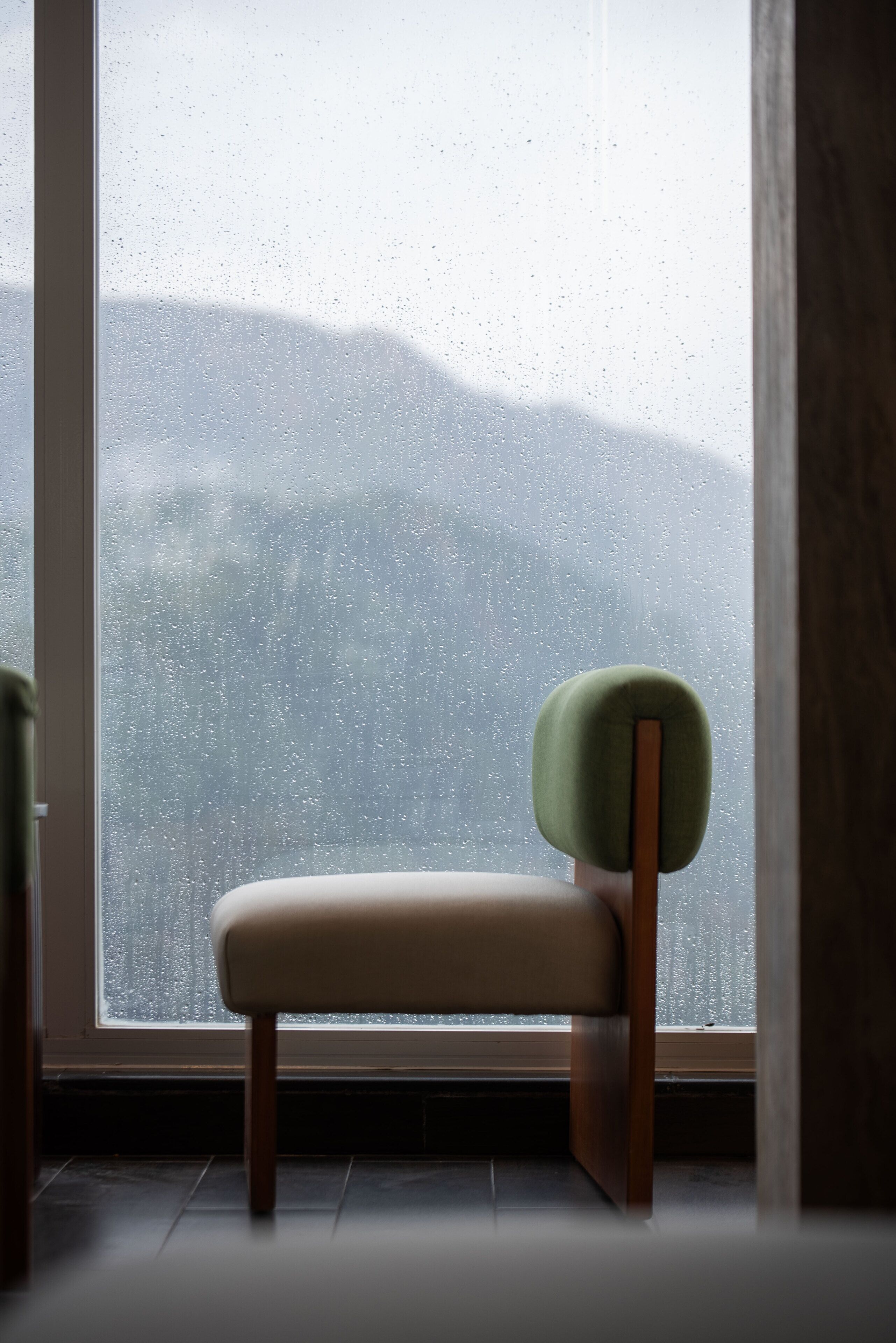 Una silla con reposacabezas verde y patas de madera, ubicada frente a una ventana con vistas a una cadena montañosa.