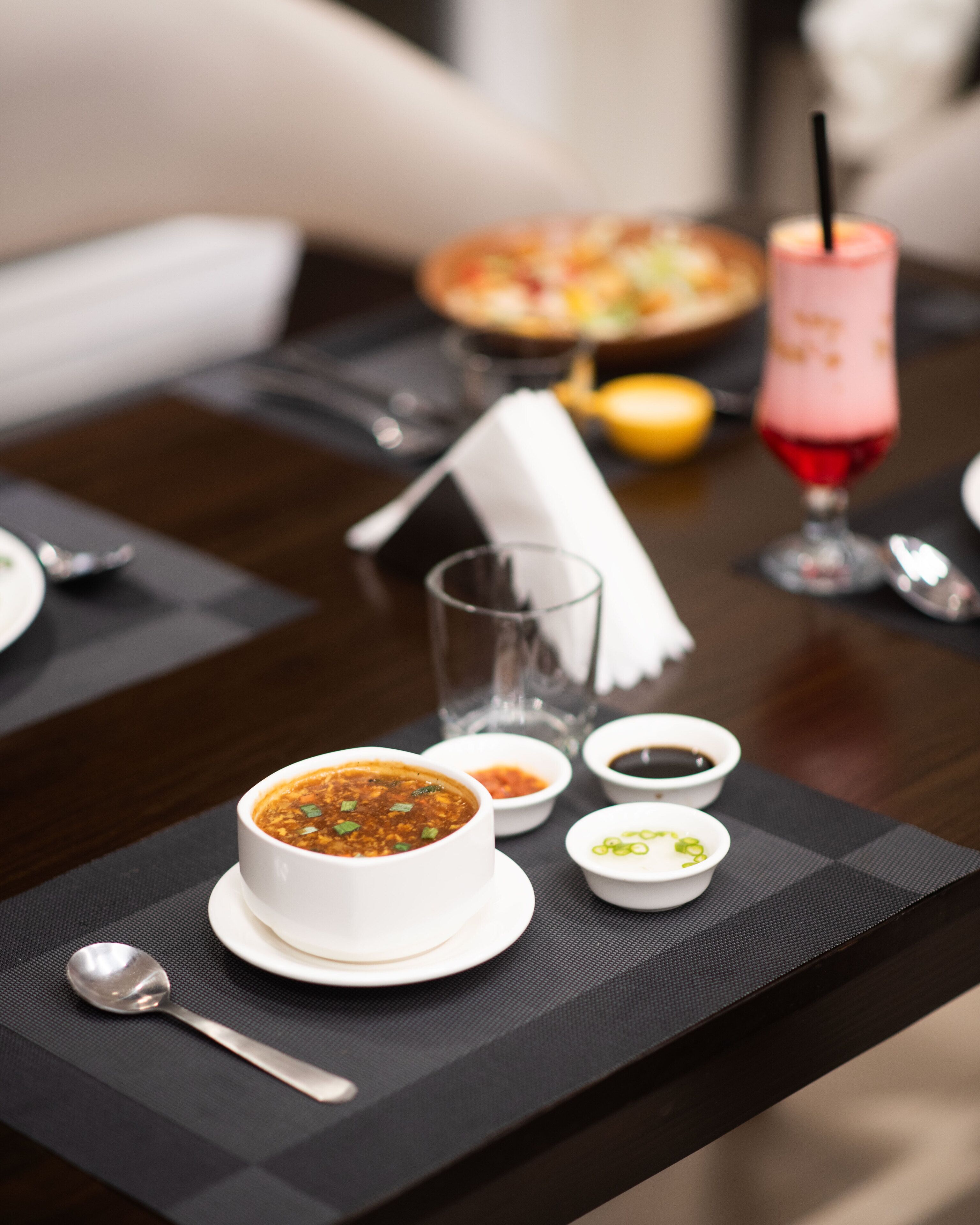 Una mesa de comedor con un plato de sopa, dos recipientes pequeños de condimentos, un vaso y una bebida rosa con pajita.
