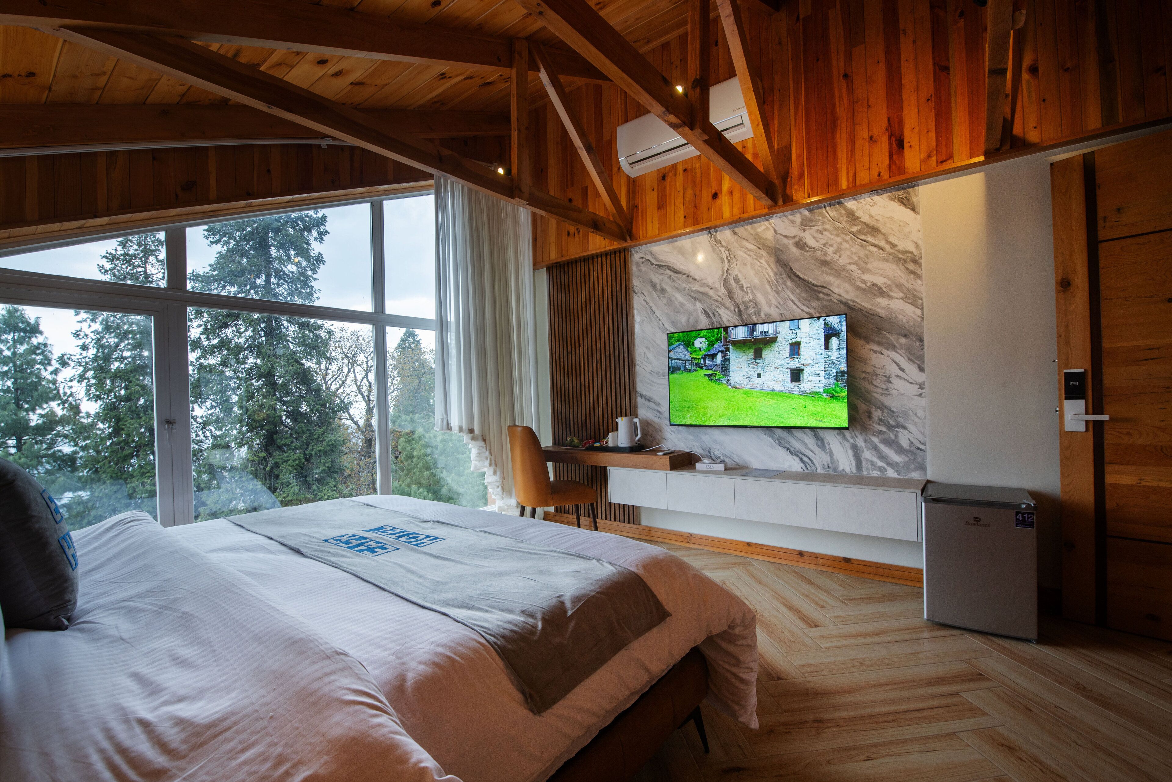 A bedroom with a large bed, a TV mounted on the wall, a small refrigerator, and a window overlooking trees.