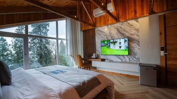 A bedroom with a large bed, a TV mounted on the wall, a small refrigerator, and a window overlooking trees.