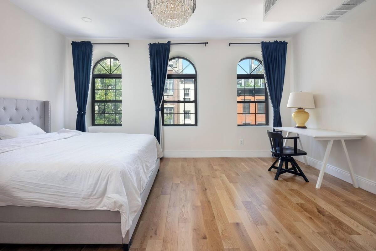 A bedroom with a bed, a desk, and three windows with curtains.