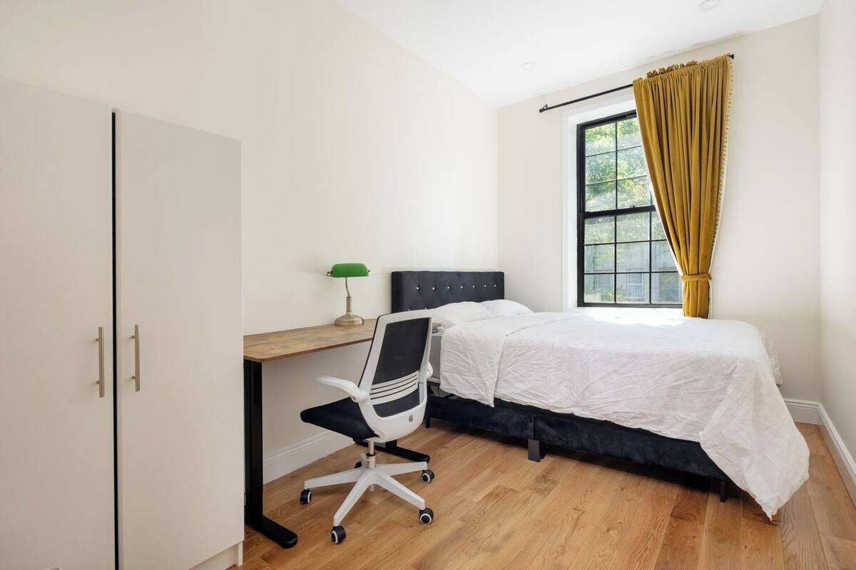 A bedroom with a bed, desk, chair, and a window with curtains.
