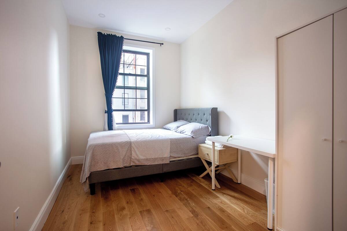 A bedroom with a bed, a bedside table, a desk, a window with curtains, and a wardrobe.