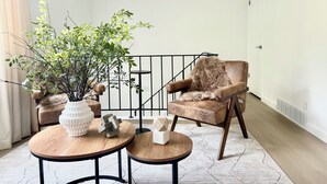 Un salon moderne avec un fauteuil en cuir marron, deux tables en bois ornées d’objets décoratifs, une plante en pot et un escalier doté d’une rampe noire.