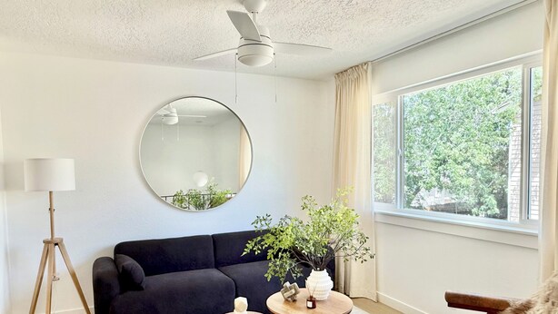 Un salon moderne avec un canapé noir, un miroir rond, un ventilateur de plafond, une grande fenêtre donnant sur un paysage verdoyant et une petite table ronde agrémentée d’objets décoratifs.