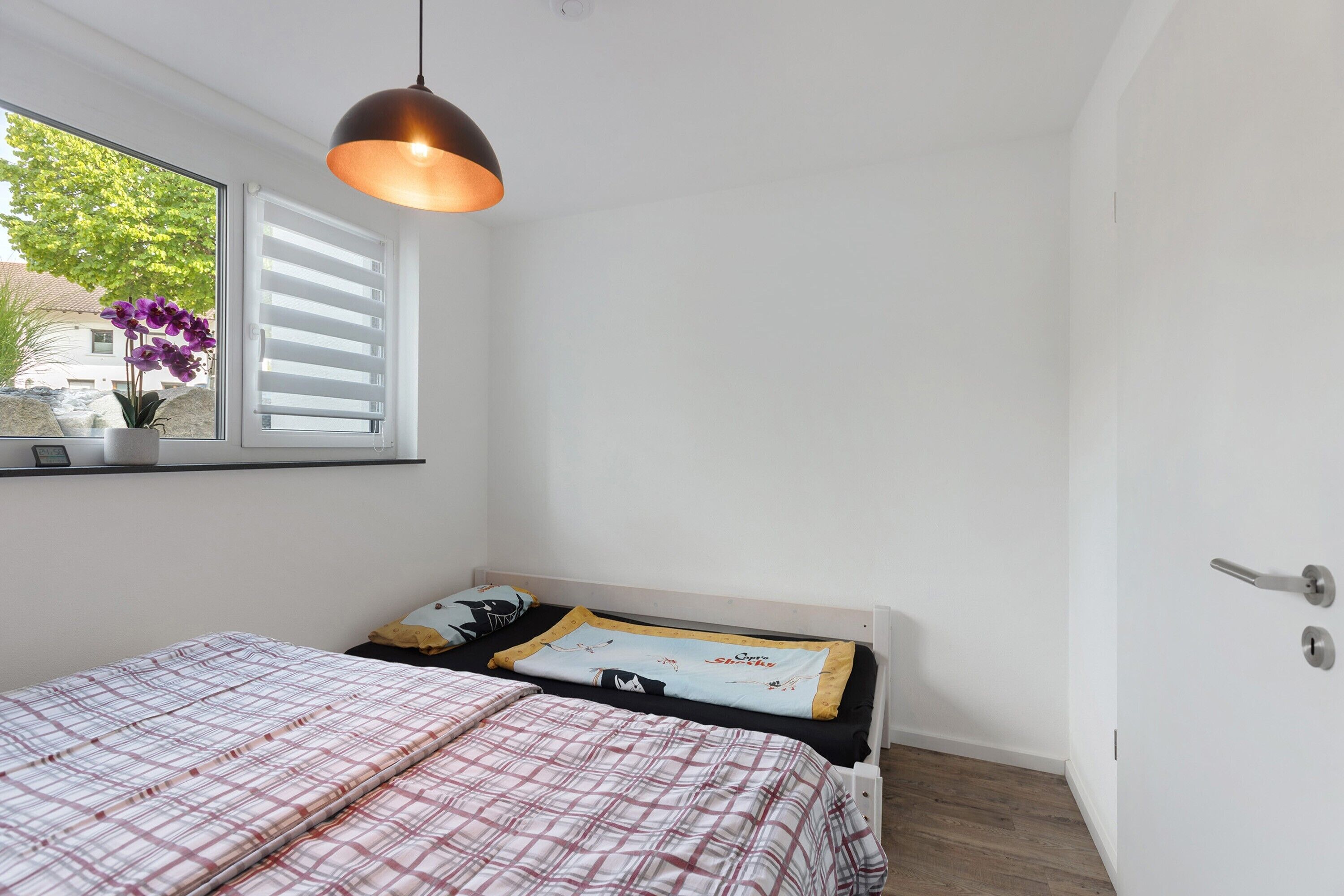 A bedroom with a bed, a window with blinds, a hanging light fixture, and a door.