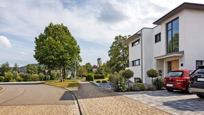 Una zona residencial moderna con un camino pavimentado, un coche rojo estacionado junto a la casa y un árbol grande en primer plano.