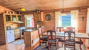 A cozy cabin interior with wooden walls, a kitchen with a refrigerator, a dining area with a table and chairs, and a window overlooking a scenic view. - Tussebu by Interhome (Tysnes)