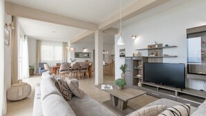 Een moderne woonkamer met een grote flatscreen televisie, een bank, een salontafel en een eethoek op de achtergrond.