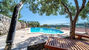 En swimmingpool med en stenmur och ett trädäck, omgiven av träd och med en fri utsikt över havet.