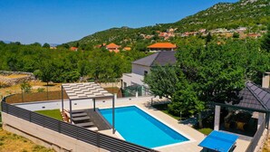 A modern pool area with a rectangular pool, a covered seating area, and a view of a hillside village.