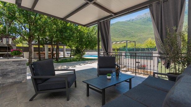 A covered outdoor seating area with a pool view, stone walls, and a pergola.