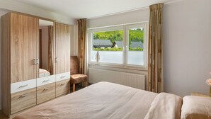 A bedroom with a bed, a wooden wardrobe, a chair, and a window with a view of greenery. - Apartment 'Dorlar' with Private Terrace, Garden, and Wi-Fi (Schmallenberg)