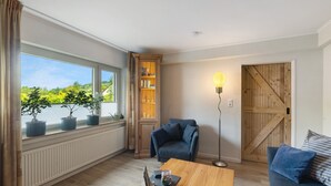 A room with a view of greenery, a bookshelf, a blue chair, a wooden coffee table, and a lamp. - Apartment 'Dorlar' with Private Terrace, Garden, and Wi-Fi (Schmallenberg)