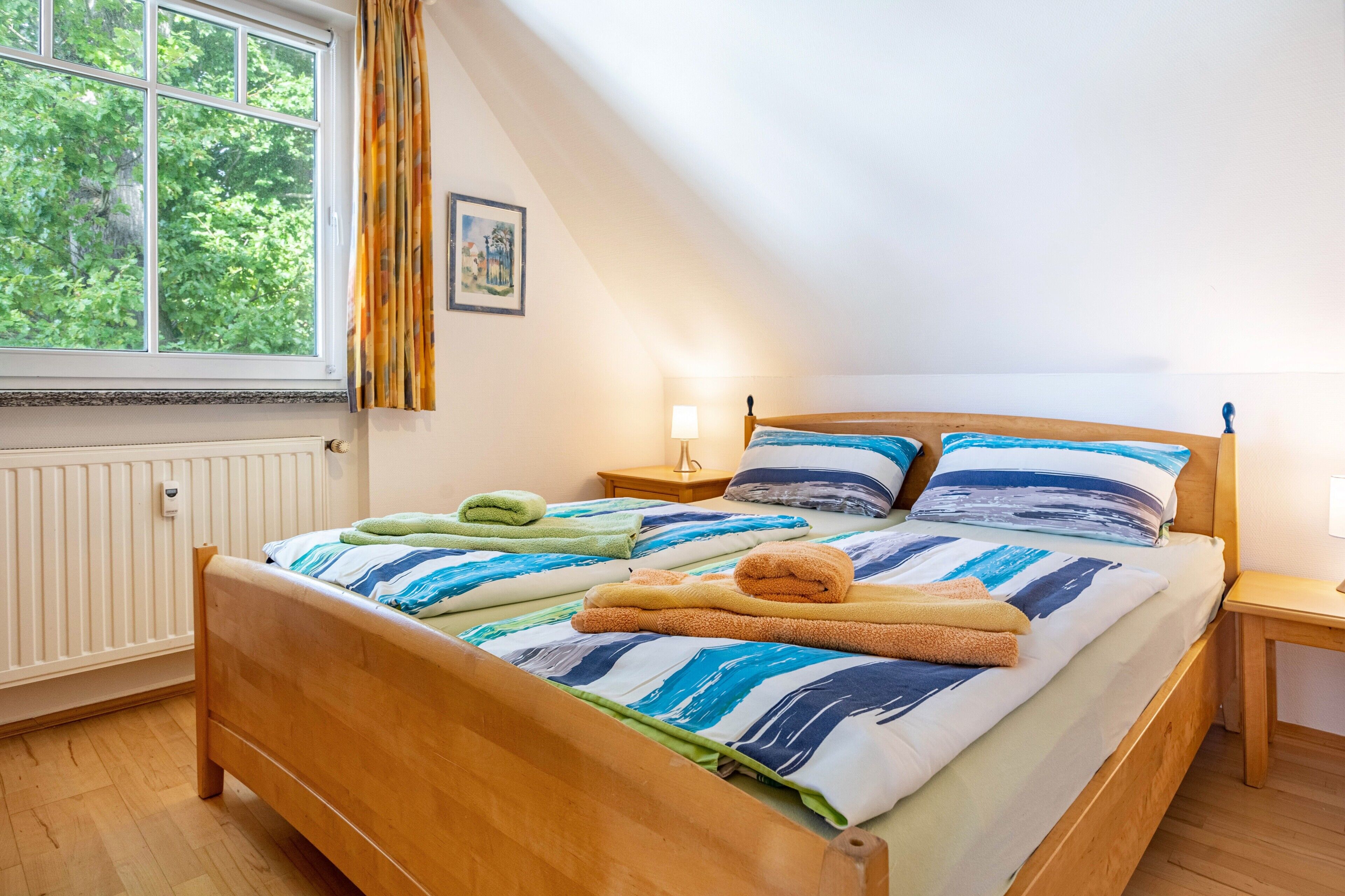 Ein Schlafzimmer mit einem hölzernen Bett, einem Fenster mit Vorhängen, einem Nachttisch mit Lampe und einem Heizkörper.