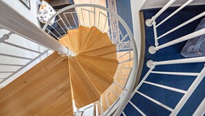 A spiral staircase with a wooden handrail and white balusters, leading to a room with a blue carpet and a patterned wall. - Apartment '22' with Balcony and Wi-Fi (Sellin)