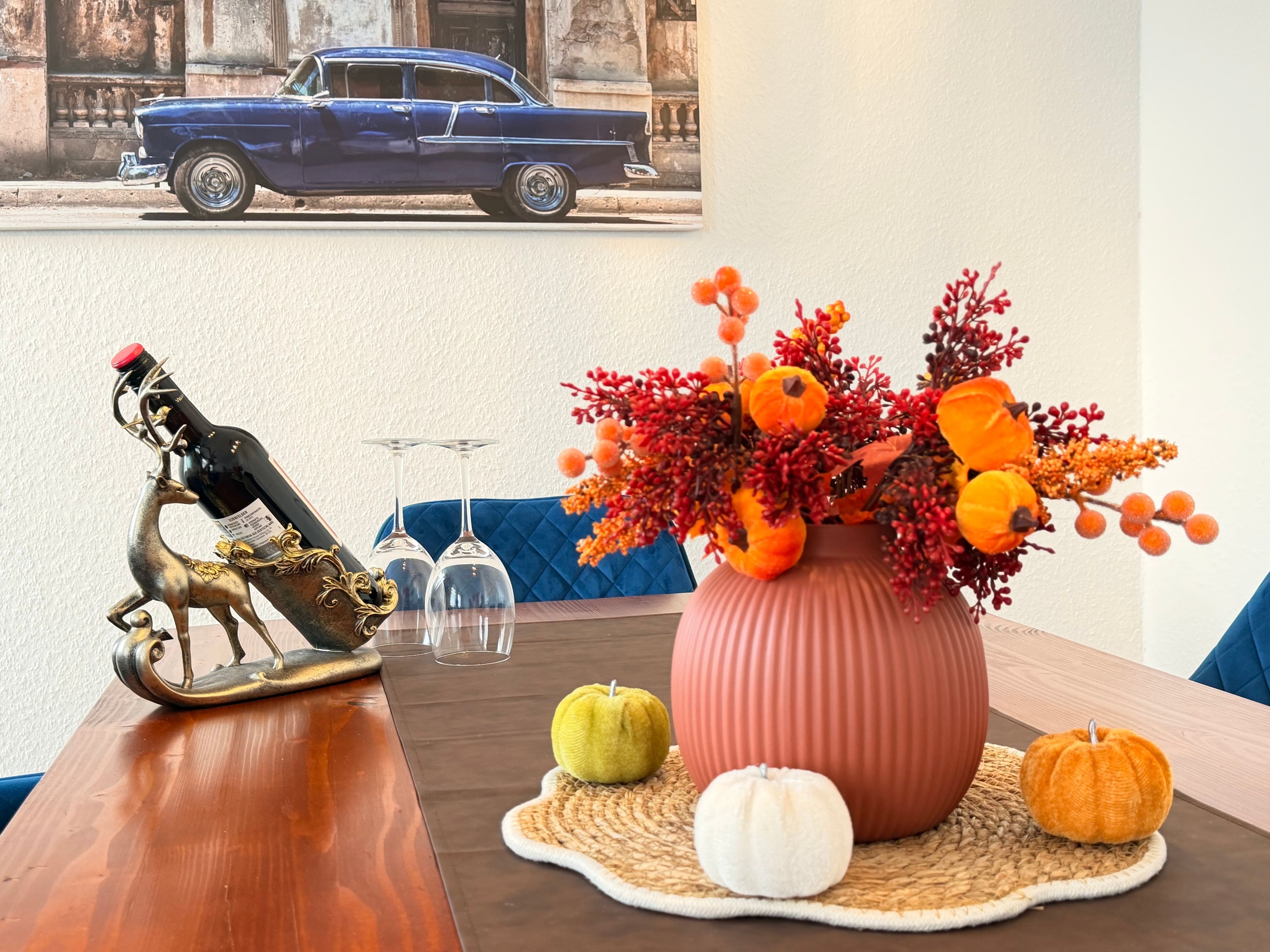 Ein Esstisch mit einer dekorativen Flasche, zwei Weingläsern, einer Vase mit Herbstblumen und einem Oldtimer im Hintergrund.