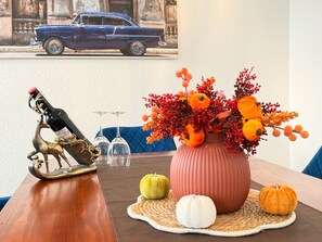 Une table à manger agrémentée d’une bouteille décorative, de deux verres à vin, d’un vase rempli de fleurs d’automne et, en arrière-plan, d’une voiture ancienne.