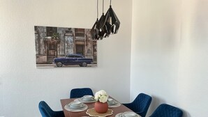 Une salle à manger avec une table en bois dressée pour quatre personnes, des chaises à l’assise rembourrée en tissu bleu, un vase avec des fleurs blanches et un tableau représentant une voiture ancienne accroché au mur.