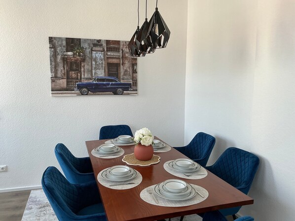 A dining room with a wooden table set for four, blue upholstered chairs, a vase with white flowers, and a wall-mounted painting of a vintage car.