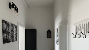 Un couloir avec une porte à gauche, un réfrigérateur de couleur sombre au centre et une porte à droite. Des luminaires sont fixés au mur et une petite plante se trouve près de la porte de droite.