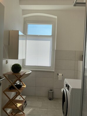 Une salle de bain moderne et compacte, dotée d’une fenêtre, d’un lavabo, d’une petite plante, d’un étendoir, d’une poubelle et d’un lave-linge.