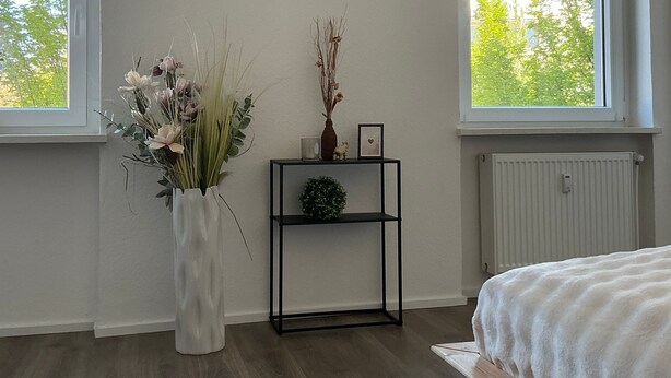 Une chambre avec une grande fenêtre, un vase blanc orné de fleurs, une étagère noire avec des objets décoratifs et un lit recouvert d’une couverture blanche.
