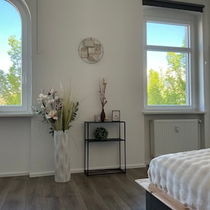 A bedroom with a large window, a white vase with flowers, a black shelf with decorative items, and a bed with a white blanket.
