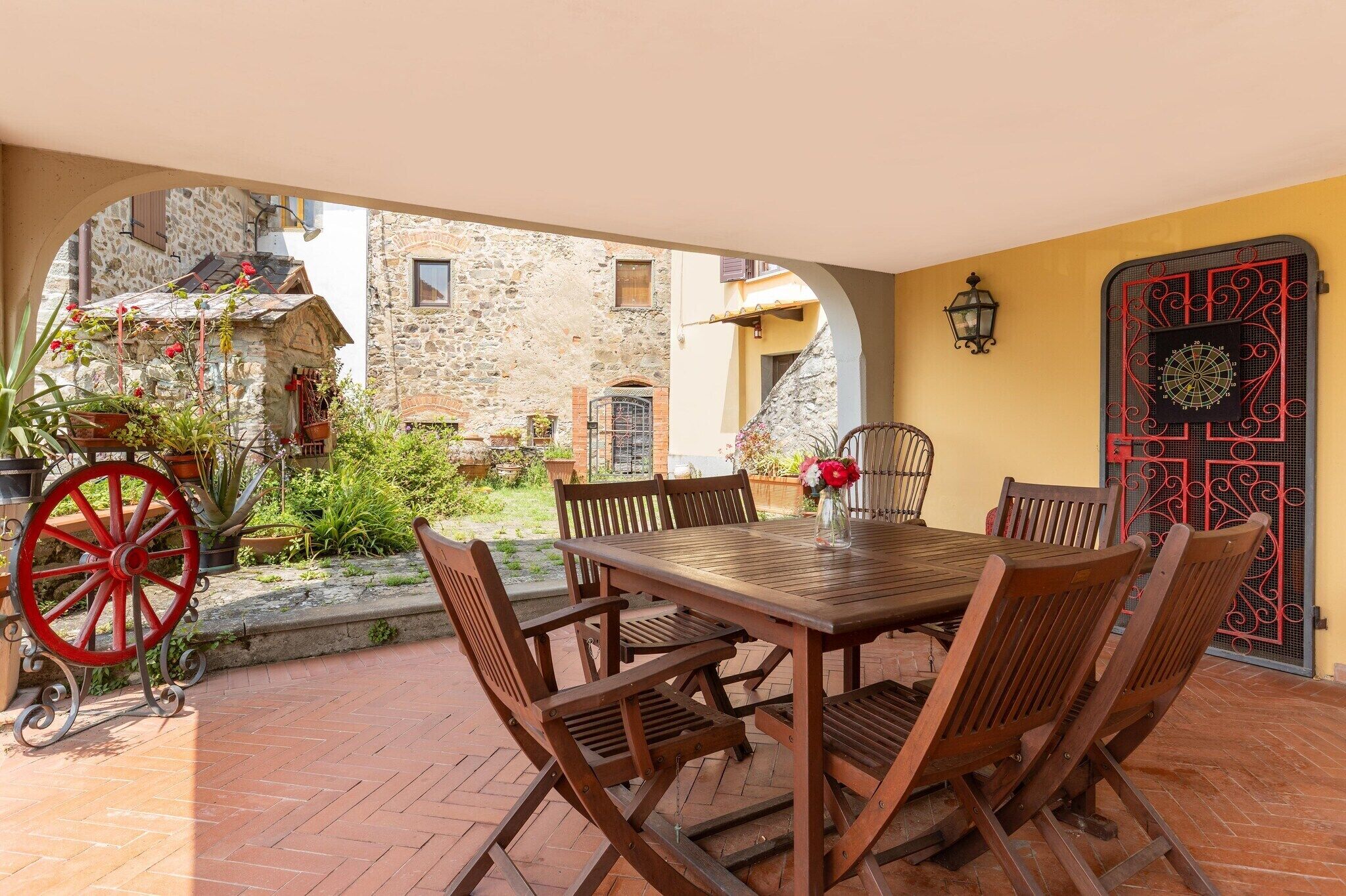 Un patio esterno con tavolo e sedie in legno, una ruota decorativa rossa e vista su edifici in pietra.
