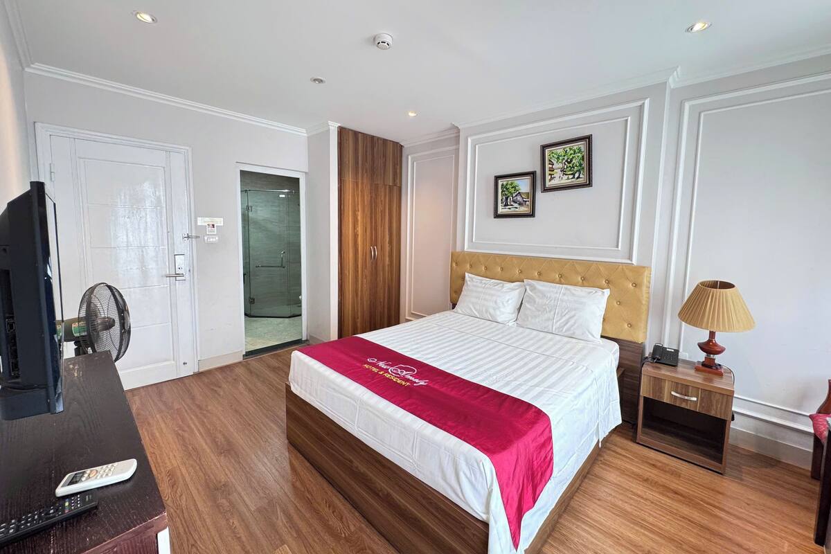 A hotel room with a large bed, a wooden wardrobe, a desk with a computer and a fan, a nightstand with a lamp, and a door leading to a bathroom.