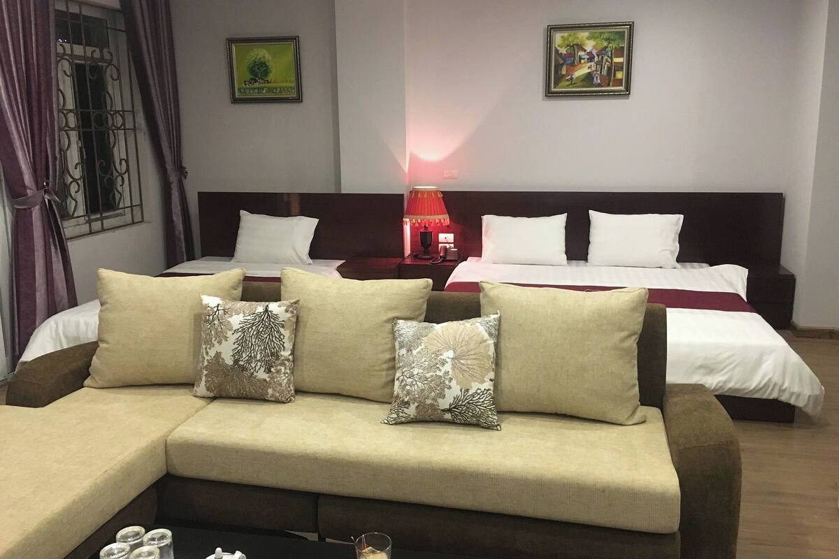 A hotel room with a sofa, two beds, a red lamp, and two framed pictures on the wall.