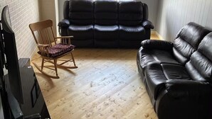 A living room with a black leather sofa, a wooden rocking chair, and a television on a stand. - Nan’s Place Vacation Home! Traditional salt box home w/ accessibility ramp (Grand Bank)