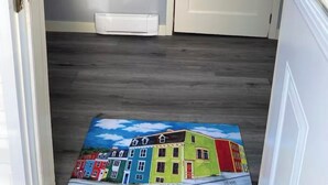 A hallway with a white door, a shoe rack with hangers, a small shelf with a coffee cup, and a colorful rug depicting houses. - Nan’s Place Vacation Home! Traditional salt box home w/ accessibility ramp (Grand Bank)