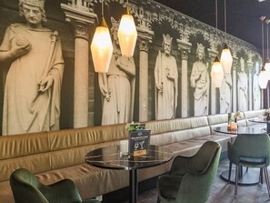 Relax in a stylish seating area with a long bench and cushioned chairs, surrounded by a beautiful mural of classical statues.