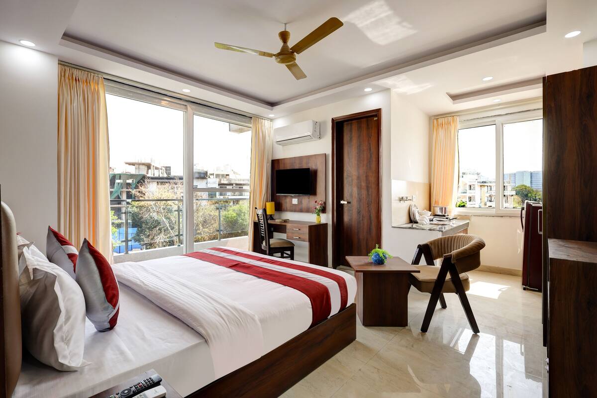 A hotel room with a large bed, a desk, a chair, a TV, and a balcony with a view of the city.