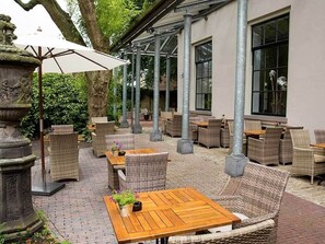 Relax in style with comfortable wicker chairs and tables under a spacious umbrella, surrounded by a columned structure and beautifully paved area.
