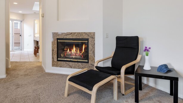 A living room with a fireplace, a black chair, a small table with a vase and a cap, and a hallway leading to a kitchen.