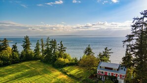 A house with a red exterior and a blue roof, surrounded by a lush green landscape and a body of water in the background. - Maxwelton Aerie | Bluffside Retreat w/ Sweeping Puget Sound Views + Firepit (Clinton)