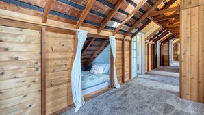 A wooden cabin interior with a high ceiling, a bedroom with a bed and curtains, and a carpeted floor. - Maxwelton Aerie | Bluffside Retreat w/ Sweeping Puget Sound Views + Firepit (Clinton)