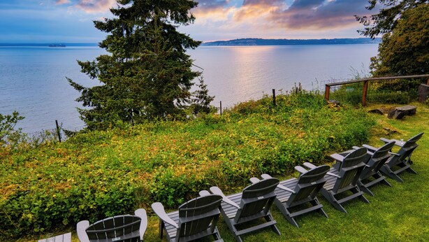A row of Adirondack chairs and a small table on a grassy area overlooking a body of water with trees and a fence in the background. - Maxwelton Aerie | Bluffside Retreat w/ Sweeping Puget Sound Views + Firepit (Clinton)